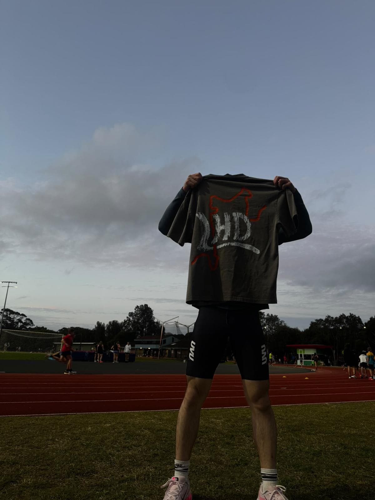 An LHD graphic T-shirt held up beside an athletics track at dusk.